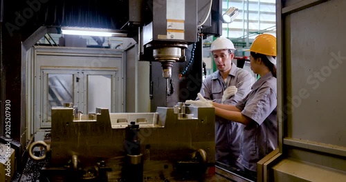 Asian man engineer briefs Asian woman technician beside CNC equipment, reviewing machining area, aligning inspection task, and supporting teamwork for precision manufacturing workflow on shop floor