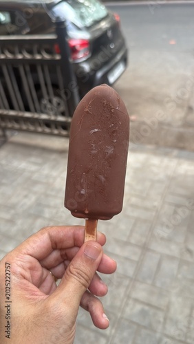 Hand-held chocolate‑coated ice cream bar (chocobar) on a stick, street background, close-up