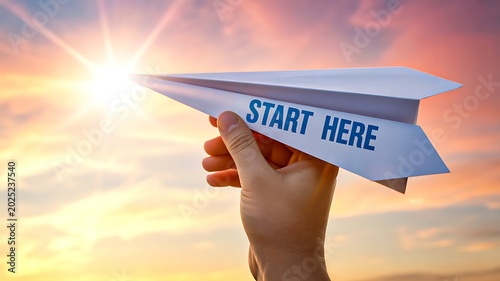 Hand holding paper airplane with start here message at sunset sky