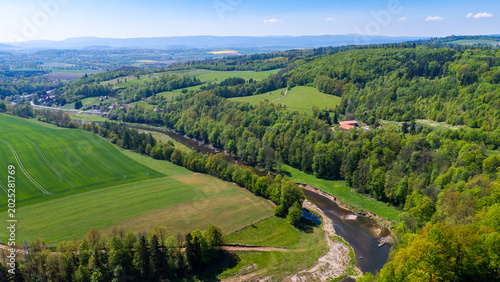 Aerial drone photograph showing the Nysa Klodzka river winding through a lush green valley in Klodzko, surrounded by dense forests, agricultural fields, and rolling hills under a bright blue sky.