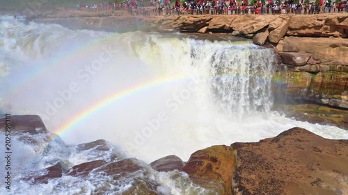 Detailed high-angle shot looking down at the crest of Hukou Waterfall. A vivid double rainbow stretches across the churning white water and rising mist as the Yellow River surges forward.