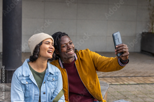 Vászonkép Couple in denim, mustard jackets sitting on concrete plaza bench taking selfie w