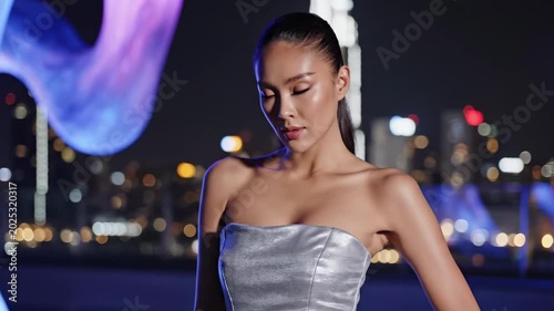 Futuristic hot thai women inspired rooftop night portrait with silver strapless dress, blue sculptural light, skyline bokeh, sleek ponytail, cinematic urban glamour style, luxury after dark fashion