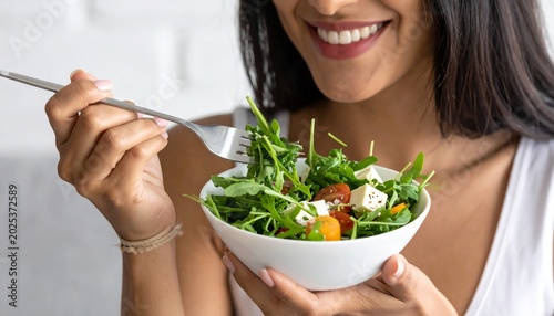 A smiling young woman eating a fresh healthy salad from a white bowl with a fork while focusing on mindful nutrition and daily wellbeing habits.