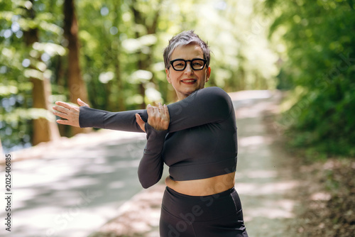 Photos Mature woman in black athletic wear stretches her arm while standing on a tree-l