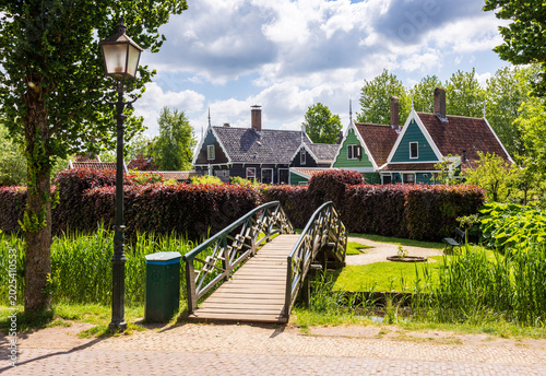 A wooden footbridge leads to the Overtuin garden lined with historic wooden houses in the Zaanse Schans quarter in Zaandam, Netherlands, on a sunny spring day.