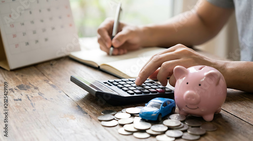 Person using a calculator and writing in a notebook to plan a budget for a new car with a piggy bank and coins on a wooden desk