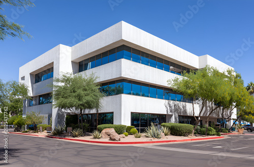 Modern three-story building with blue-tinted windows, lush greenery, and desert landscaping under a clear, bright, blue sky