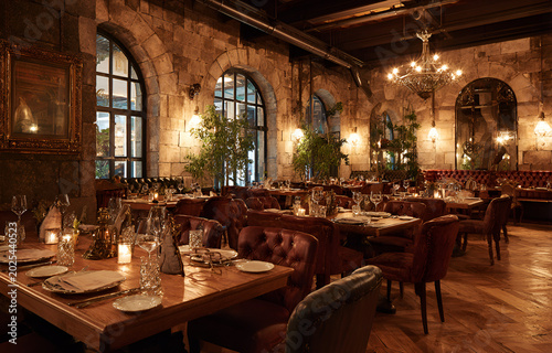 Interior restaurant view with brick walls, arched windows, wooden tables, and velvet seating under dim lighting. Chandeliers hang above