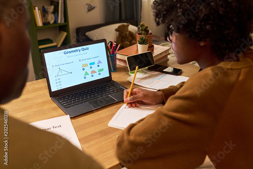 Obraz na plátně Black teenager studying geometry on laptop, taking notes during online tutoring session at home with parent nearby