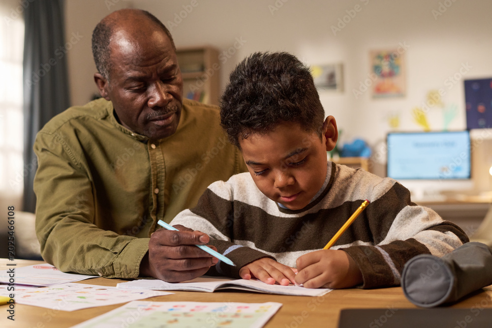 Naklejka premium Black senior man assisting schoolboy with homework, guiding writing practice at home and supporting learning progress. Useful for parenting, tutoring, education campaigns, medium shot