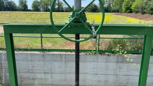 Water Gate Mechanism: A sturdy green metal water gate mechanism, featuring a large wheel and chain, stands beside a concrete wall, overlooking fields in the distance under a clear blue sky.