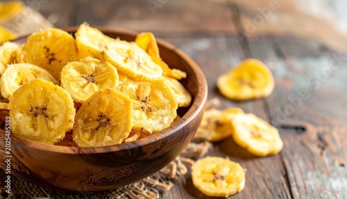 A bowl of banana chips sits on a rustic wooden table surrounded by scattered slices.