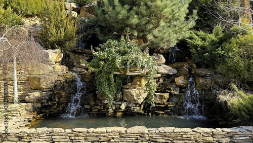 Rocky garden waterfall cascading into pond with layered stone wall, moss covered boulders, evergreen shrubs, sunlit reflections,