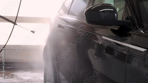 Car wash, high pressure water jet rinsing foam from black car at indoor station. Strong spray clears dirt, dynamic cleaning, automotive service and vehicle care