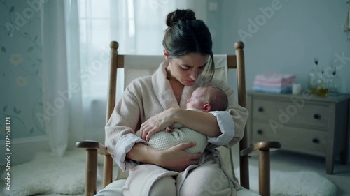 Cradling woman in robe fixing swaddle and rocking chair in nursery as infant stirs, checking breath