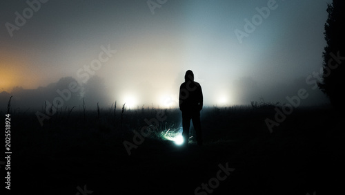 A mysterious hooded figure standing next to a glowing light in a field on a spooky foggy night