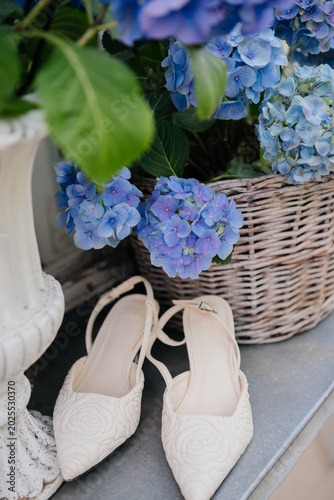 Fotografía A pair of chic white slingback bridal shoes with textured embroidery patterns placed near a rustic wicker basket filled with blooming blue hydrangea flowers