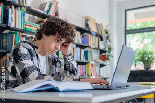 Fotografía Teenage boy looking bored while studying on laptop in library