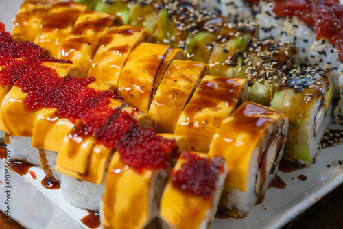 A vibrant platter of assorted fusion sushi rolls topped with melted cheese, bright red tobiko, sliced avocado, and a savory drizzle of unagi sauce.