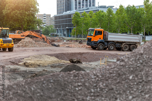 Foto Urban construction site with excavator and dump truck during sunrise near modern buildings and green trees