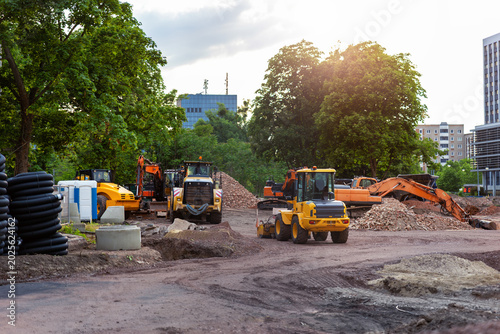 фотографія Urban construction site with excavator and dump truck during sunrise near modern buildings and green trees