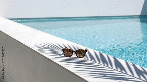 Tortoiseshell Sunglasses on White Ledge of Luxury Swimming Pool with Palm Leaf Shadow Minimalist Summer Vacation Concept