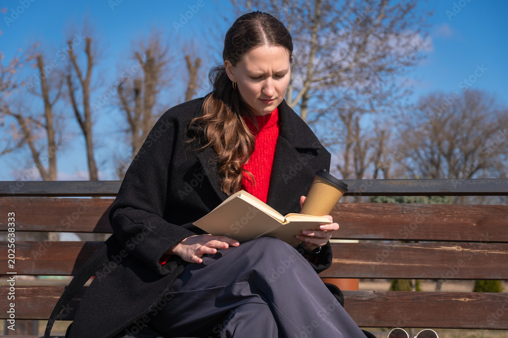 Obraz premium Young woman on a wooden park bench reading a book and holding a takeaway coffee, enjoying a quiet, thoughtful moment outdoors in cool weather among bare trees and sunlight