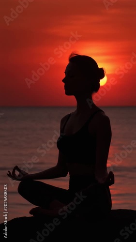 Silhouette of a woman meditating in lotus pose by the sea at sunset, with a vibrant red and orange sky reflecting on the water.