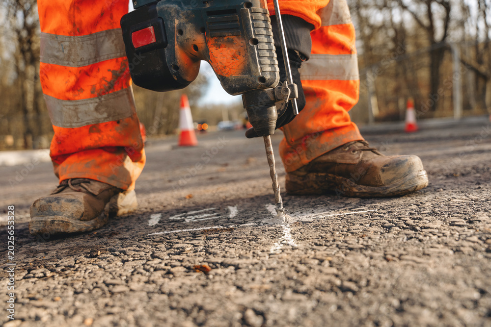 Obraz premium Worker drills into asphalt at a road construction site in the late afternoon near a busy area with traffic