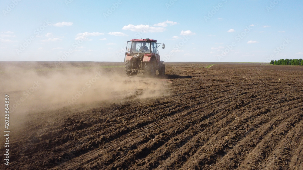 Fototapeta premium drone shot capturing a red tractor pulling a harrow across a vast, dark-soiled field. Dust trails behind the machinery as it prepares the land for the planting season under a clear blue sky.