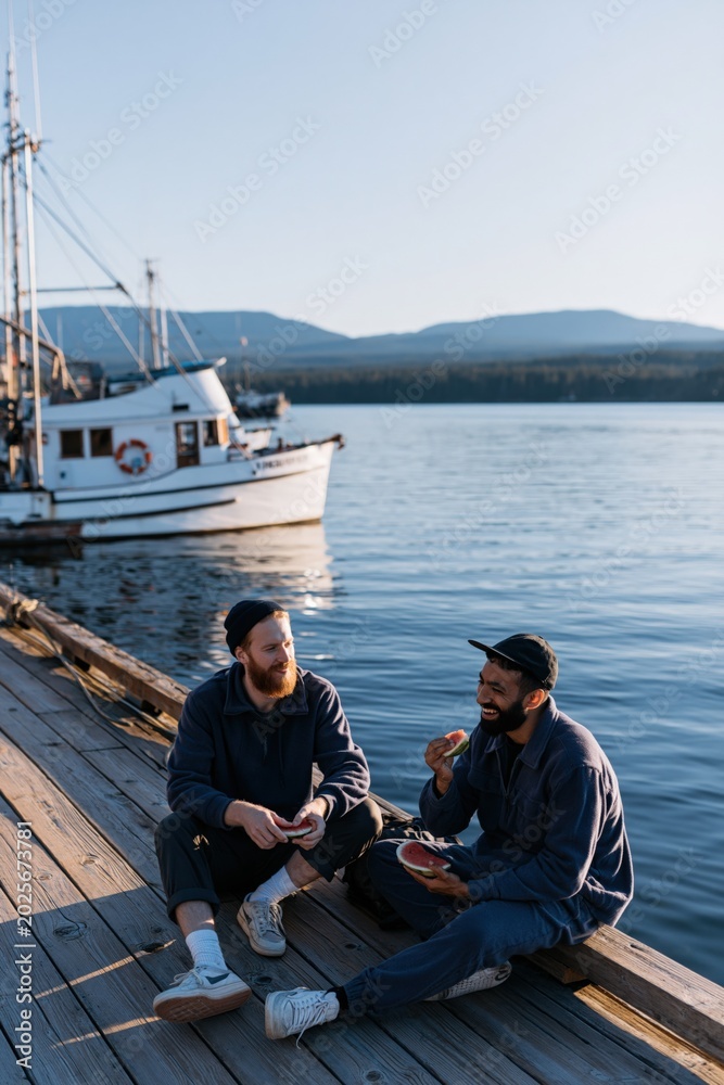 Fototapeta premium Two men laughing and eating on a wooden dock near a fishing boat for lifestyle banner and coastal background