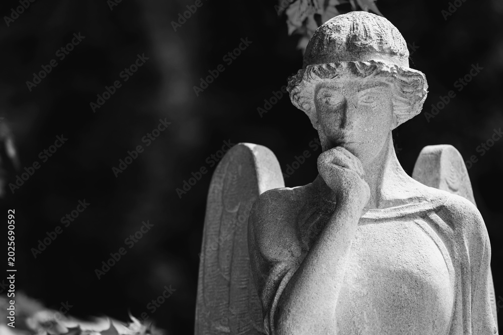 Fototapeta premium Monochrome angel statue with thoughtful expression in dramatic cemetery atmosphere