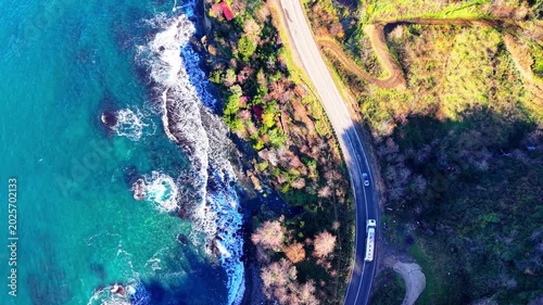 Bird’s-eye drone footage tracking vehicles as they move along an asphalt road between dense green forest and crystal-clear turquoise sea. The camera remains pointed straight down while flying forward,