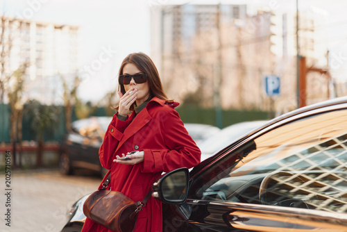 Foto Stylish woman in a red coat stands beside a black car, covering her mouth with her hand, in an urban setting with tall buildings and parked cars
