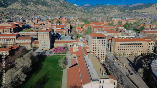 Lawn and foothill alignment near Broadway with academic halls, interior courtyards, walkways leading toward foothill edge neighborhoods. Stone facade, red roofs, scattered trees, Boulder, CO