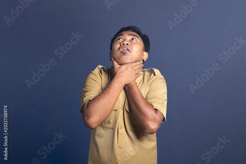 Photography Panicked young man in a khaki uniform shirt clutching his neck with both hands in a choking gesture while looking upward with open mouth against a blue background