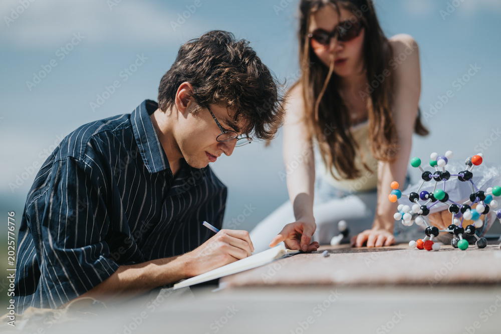 Fototapeta premium Two students study chemistry outdoors with a molecular model and notebook. A young man writes notes while his classmate points and helps nearby.
