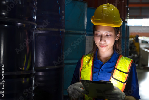 Asian female worker with tablet inspect product in large steel drums at factory during night shift. Engineer woman with helmet and safety vest work in industrial site, check product quality in a dark