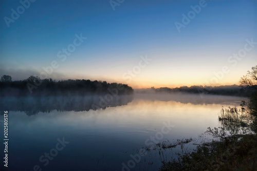 A river reflects the sky at dawn, with mist hovering over the water and trees bordering the edges. The sun begins to rise, lighting the scene softly