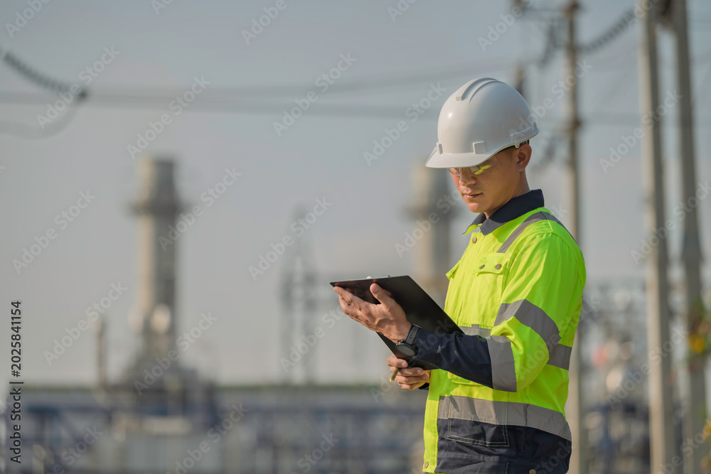 Obraz premium Asian male electrical engineer holding digital tablet standing at power plant station. Professional energy industry technician working on infrastructure maintenance and renewable resource project.