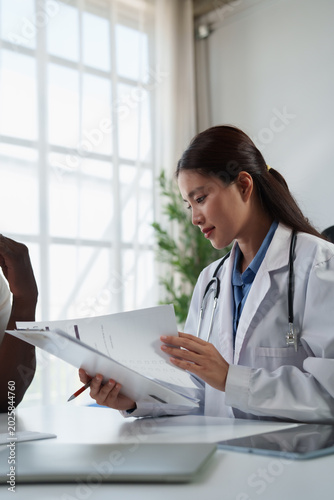 Female asian doctor having a medical consultation with a patient in a modern healthcare clinic, reviewing medical records and discussing diagnosis or treatment plan