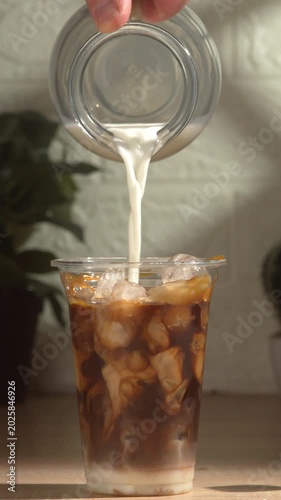 Close-up of making a takeaway iced coffee cocktail. Milk is poured from a glass jug into a plastic cup filled with layers of condensed milk, ice cubes, caramel, and coffee