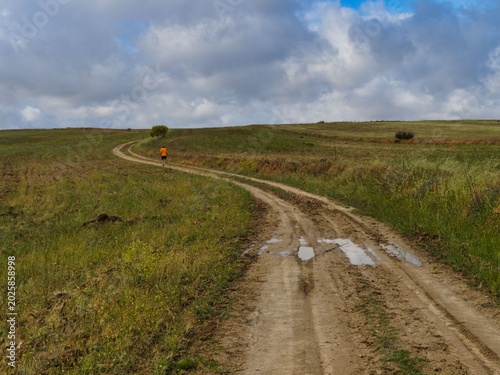 Runner moves along a winding dirt path in an open field under a cloudy sky in the afternoon sunlight