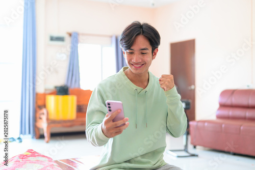 Happy young Thai man from Isan sitting using phone raising fist celebrating win