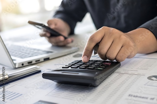 Person using a calculator while working on a laptop and holding a smartphone at a desk