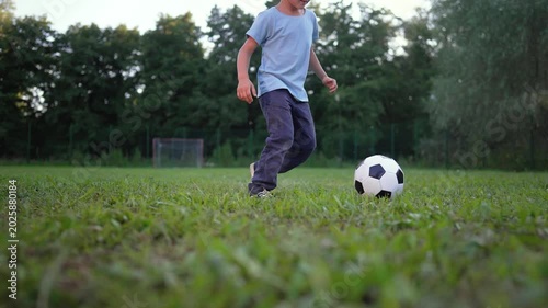 Boy runs with soccer ball on grass field at park during evening. Child plays football outdoors near goal. Kid dribbles soccer on grass. Boy with ball on field. Child running with football at park.