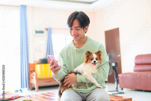Happy young Thai man from Isan sitting holding dog using phone in living room