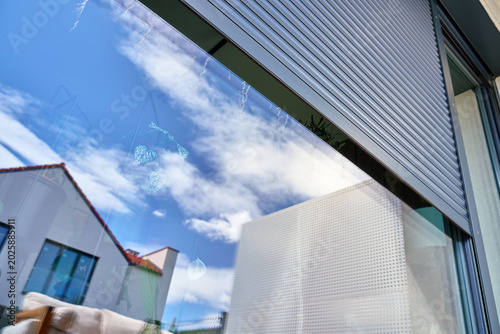 Grey exterior roller shutter is partially open above a large glass window on a modern house facade. Home security, privacy, sun protection and residential exterior