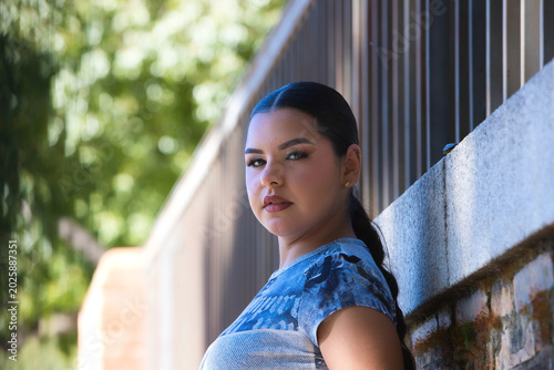 Bild auf Leinwand A young, pretty Gypsy woman from Seville, with long dark hair tied back in a ponytail and dressed in casual clothes, is leaning against a wall, smiling happily